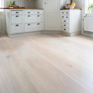 Ett ljus och öppen golvyta med breda, blekt träfärgade laminatplank som ger ett rent och enhetligt utseende i ett ljust rum med klassiska köksskåp.