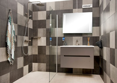 Ett stilfullt badrum med stora kvadratiska kakelplattor i svart och grått, en dusch med glasvägg, och en modern grå badrumsmöbel under en belyst spegel.