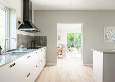 Ett modernt och ljust kök med minimalistisk design, vita skåp, en rostfri diskbänk, och en köksö som öppnar upp mot en välkomnande vardagsrumsdel.