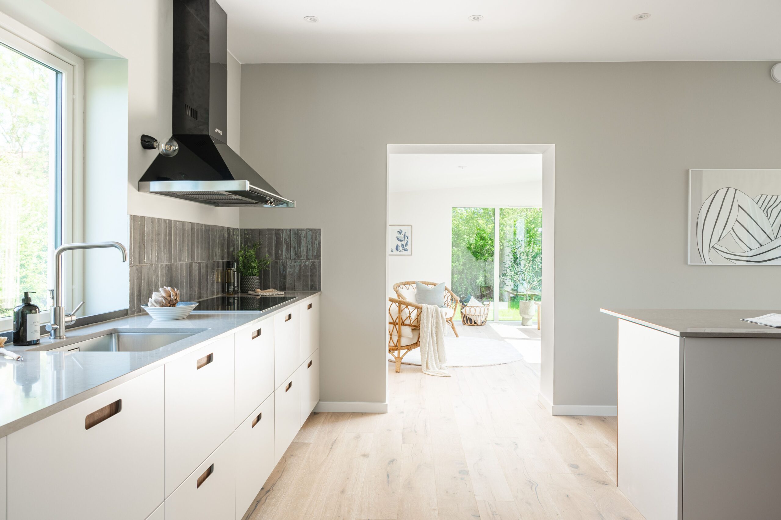 Ett modernt och ljust kök med minimalistisk design, vita skåp, en rostfri diskbänk, och en köksö som öppnar upp mot en välkomnande vardagsrumsdel.