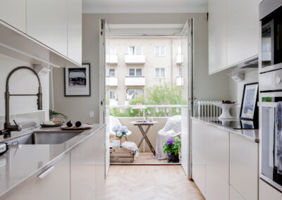 Kök i lägenhet med vita skåp och rostfria vitvaror som öppnar upp mot en mysig balkong med sittplats och gröna växter