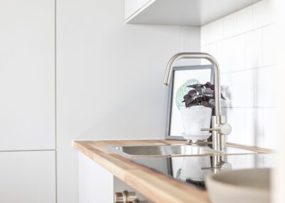 Ett ljust kök med ren design, en diskbänk i rostfritt stål och en krukväxt speglad i kranens vatten.