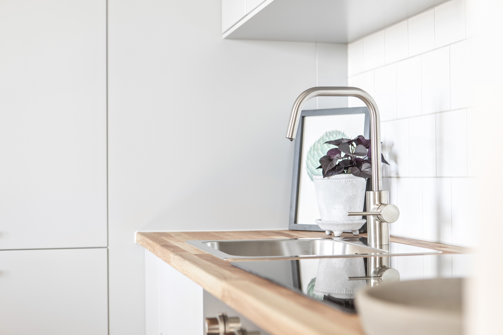 Ett ljust kök med ren design, en diskbänk i rostfritt stål och en krukväxt speglad i kranens vatten.