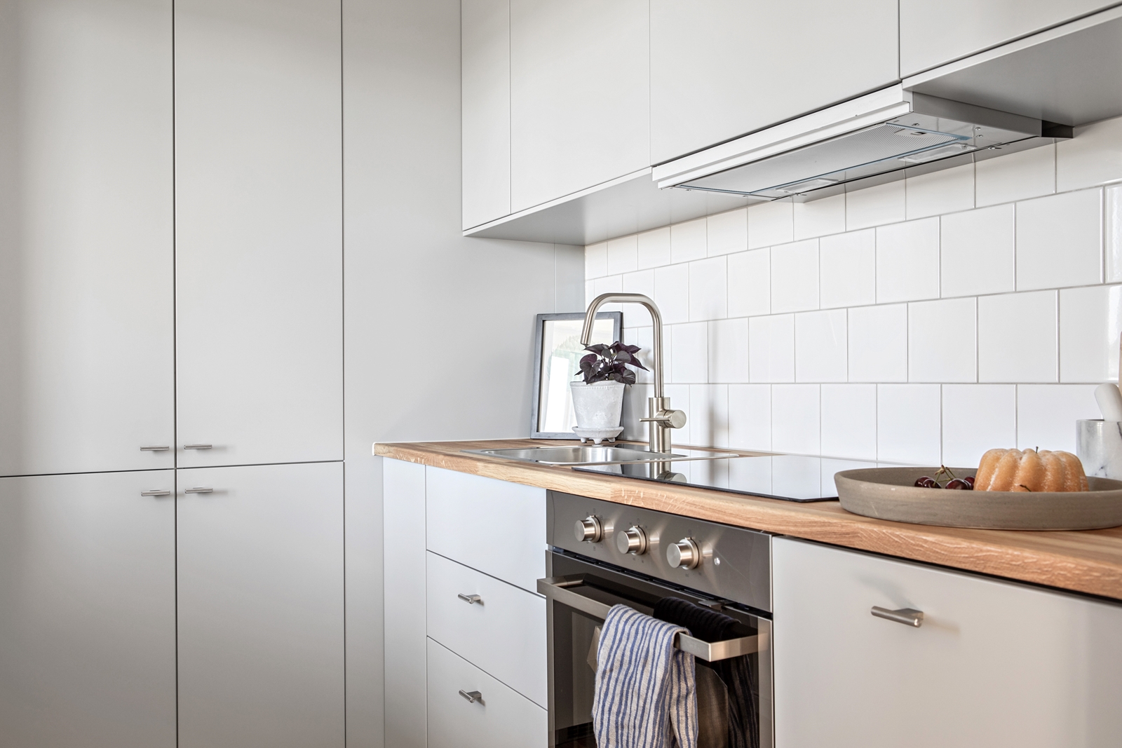 Efter en köksrenovering presenteras ett minimalistiskt kök med nya, vita, släta skåp och en diskbänk i rostfritt stål, accentuerad av en träliknande bänkskiva.