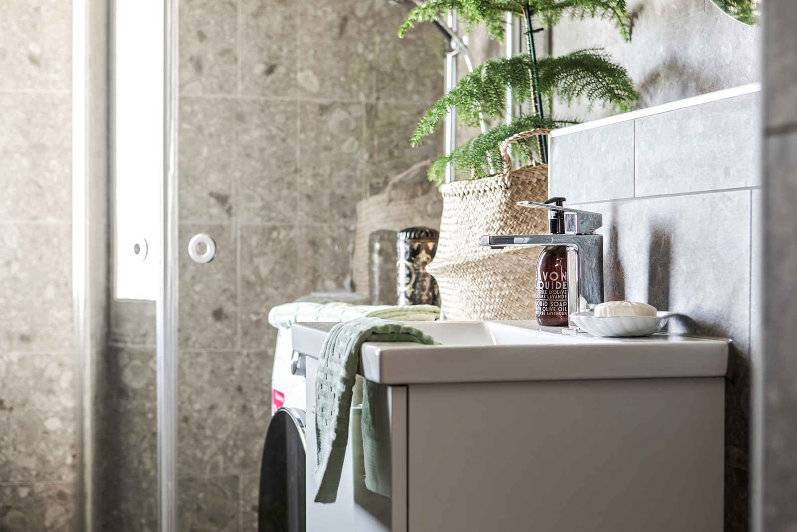 Badrum med grå marmorkakel, en modern handfat med tvål och handduk, och en korg med gröna växter som skapar en fridfull atmosfär.