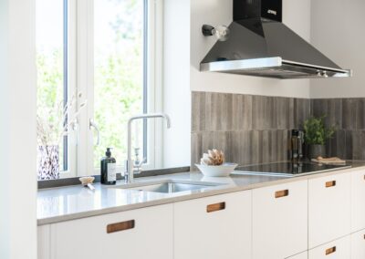 Ett kök med vita luckor, rostfria detaljer och en grå kakelvägg, belyst av naturligt ljus från ett fönster.