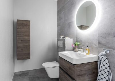 Ett nyligen renoverat badrum med stilrena gråa klinkerplattor, ett modernt handfat med trädetaljer och en rund spegel med inbyggd belysning.