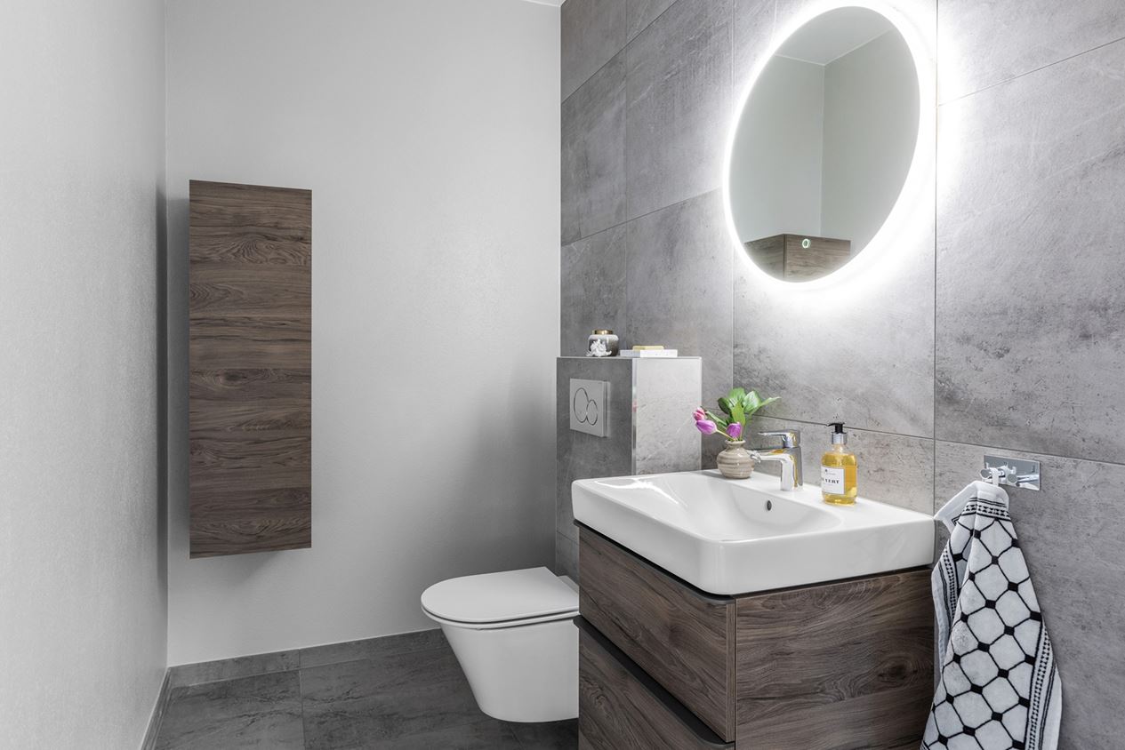 Minimalistiskt badrum med grå betongliknande kakel, rund spegel med LED-belysning och träskåpsinredning.
