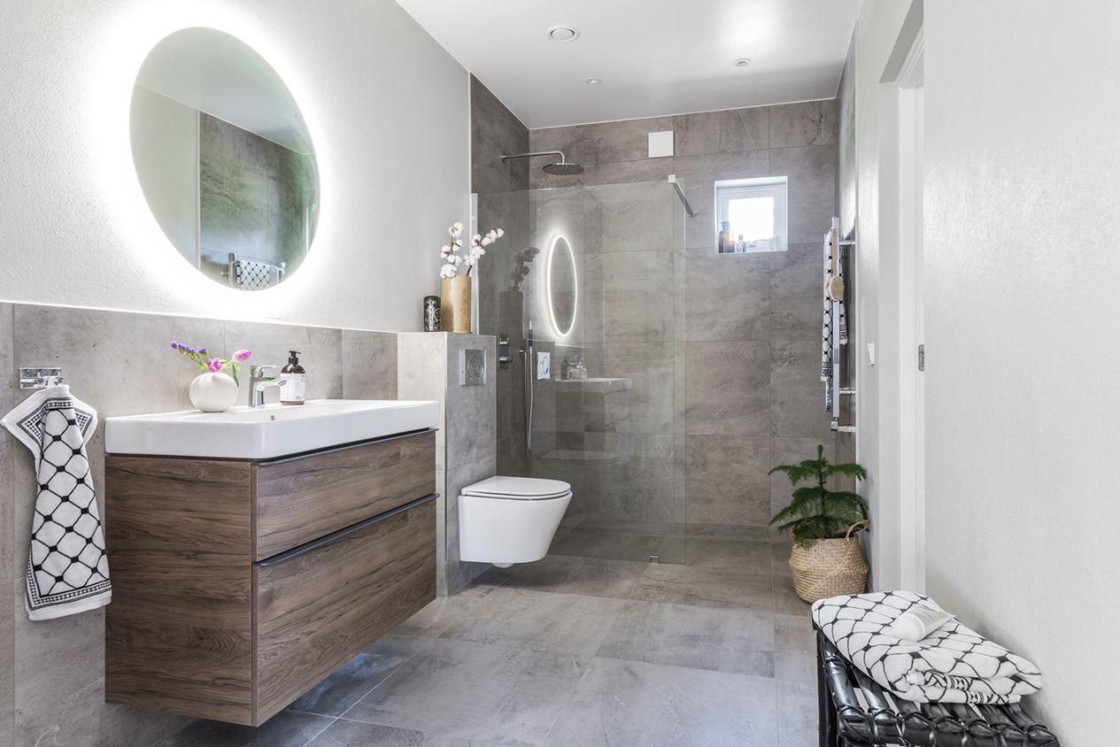 Efter en badrumsrenovering visas en modern och ljus interiör med en flytande träkommod, en rund spegel med belysning, och en genomgående grå stentextur.