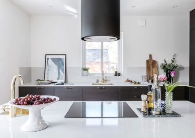 Ett stilfullt kök med mörka träskåp och en central köksö, toppad med en skinande vit bänkskiva och moderna köksredskap, belyses av en stor svart köksfläkt.