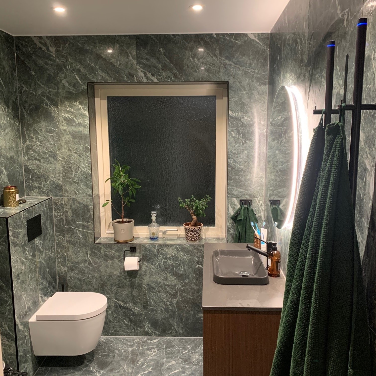Modernt badrum klätt i mörkt marmormönstrat kakel med ett fönster, ett upphängt toalett, och en elegans handfatsektion.